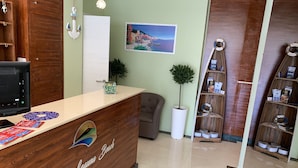 Una reception con scrivania in legno e un logo, una pianta in vaso, un quadro al muro e una mensola in legno con diversi oggetti.