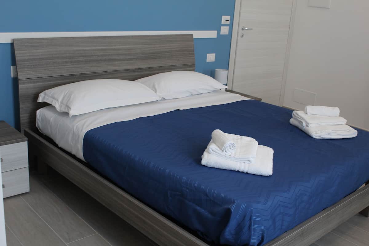 Un lit bien fait, avec une couvre-lit bleue et des oreillers blancs, une tête de lit en bois et une table de chevet blanche dotée d’un tiroir.