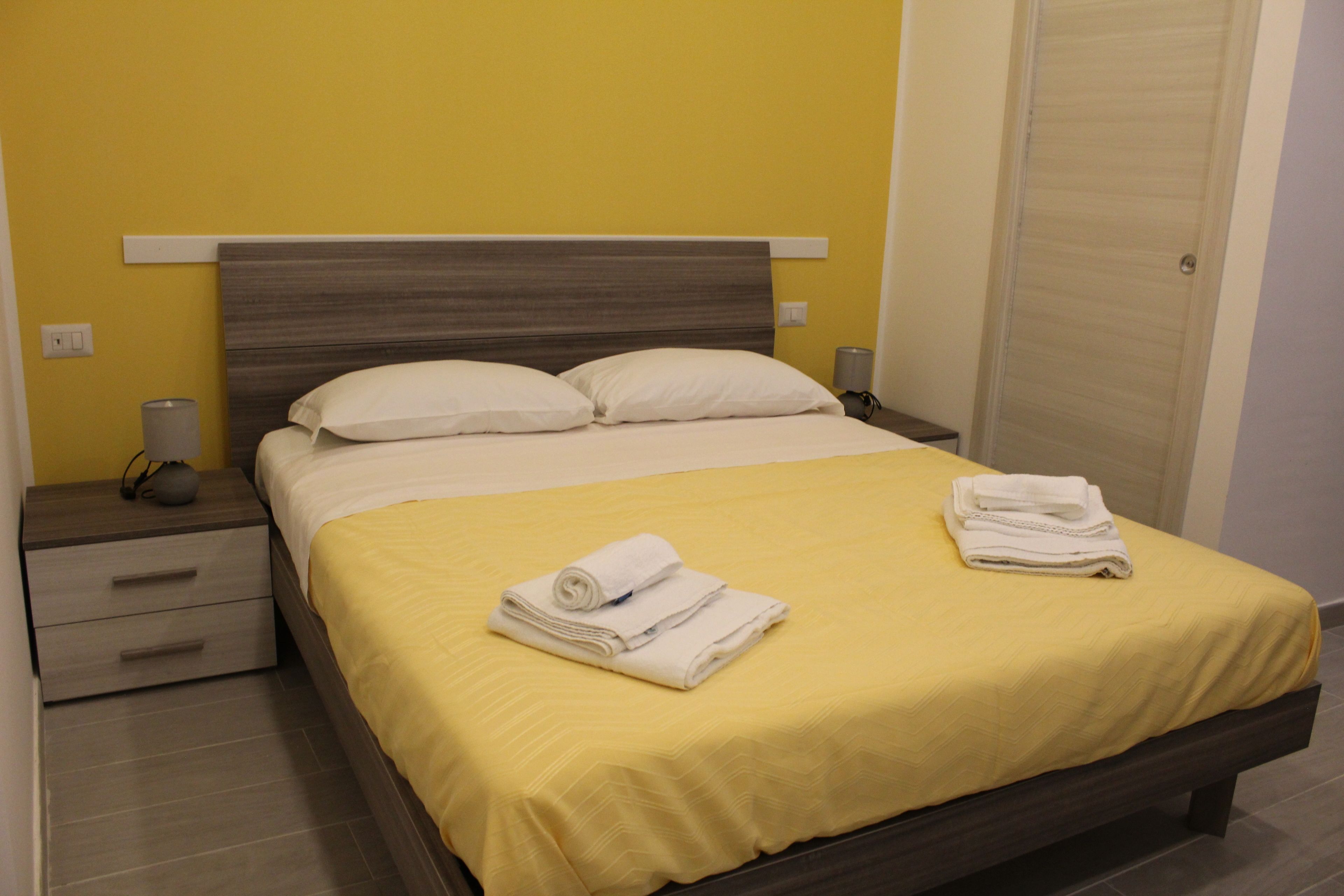 A neatly made bed with a yellow bedspread and white pillows, a bedside table with a lamp, and a closed door.
