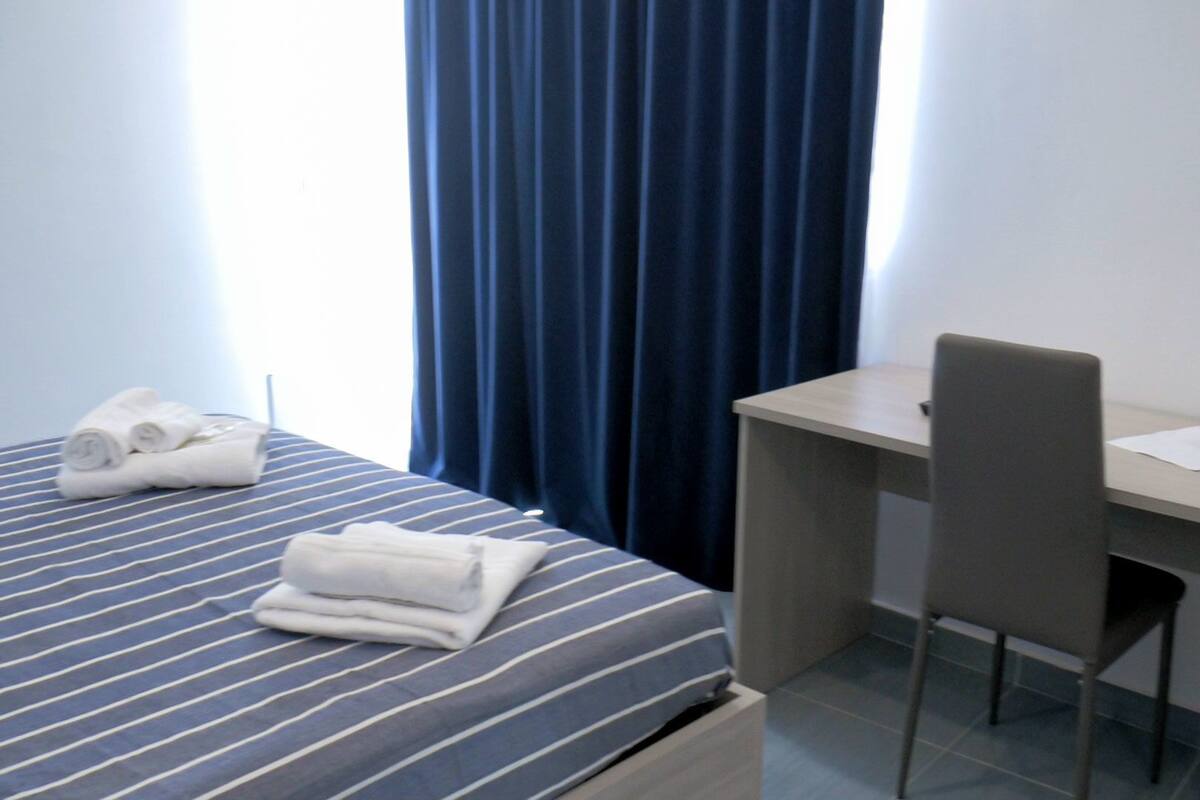 Une chambre d’hôtel avec un lit, un bureau et une chaise. Il y a des serviettes sur le lit et un vase avec une fleur sur le bureau.