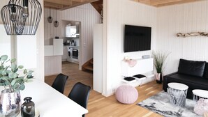 Een moderne woonkamer met een witte bank, zwarte kussens, een aan de muur bevestigde televisie en een eethoek met een witte tafel en stoelen.