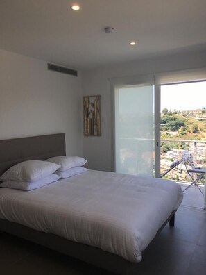 A bedroom with a large bed, a ceiling vent, a wall-mounted artwork, and a sliding glass door leading to a balcony with a view of buildings and greenery. - Twenty Rent (Cagnes-sur-Mer)