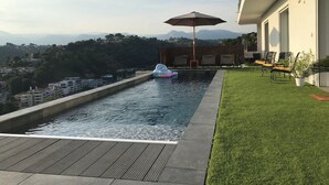 A swimming pool with a view of a hilly landscape, a lounge area with chairs, and a potted plant.