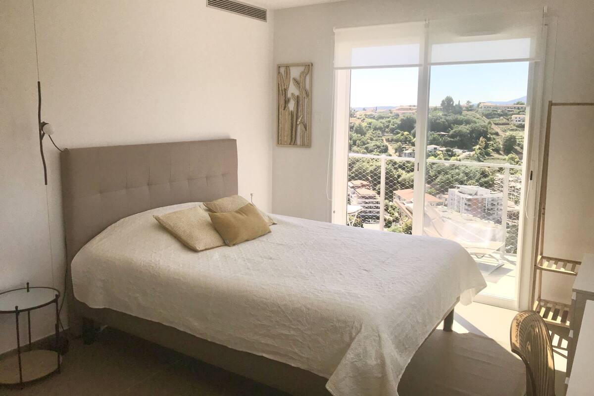 A bedroom with a large bed, a view of the city through a balcony, and a painting on the wall.