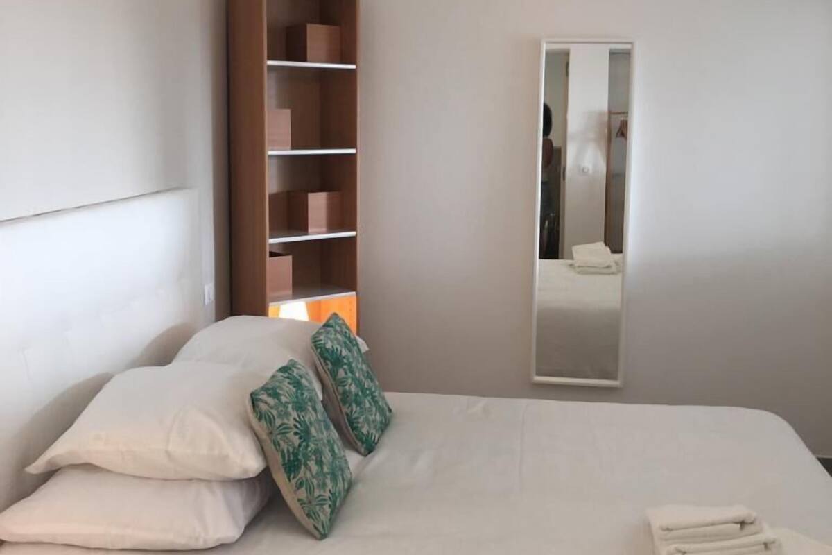 A hotel room with a bed, a wooden bookshelf, a mirror, and a hanging light fixture.