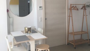 A room with a dining table, chairs, a small stool, a mirror, a wardrobe, a coat rack, and a door.