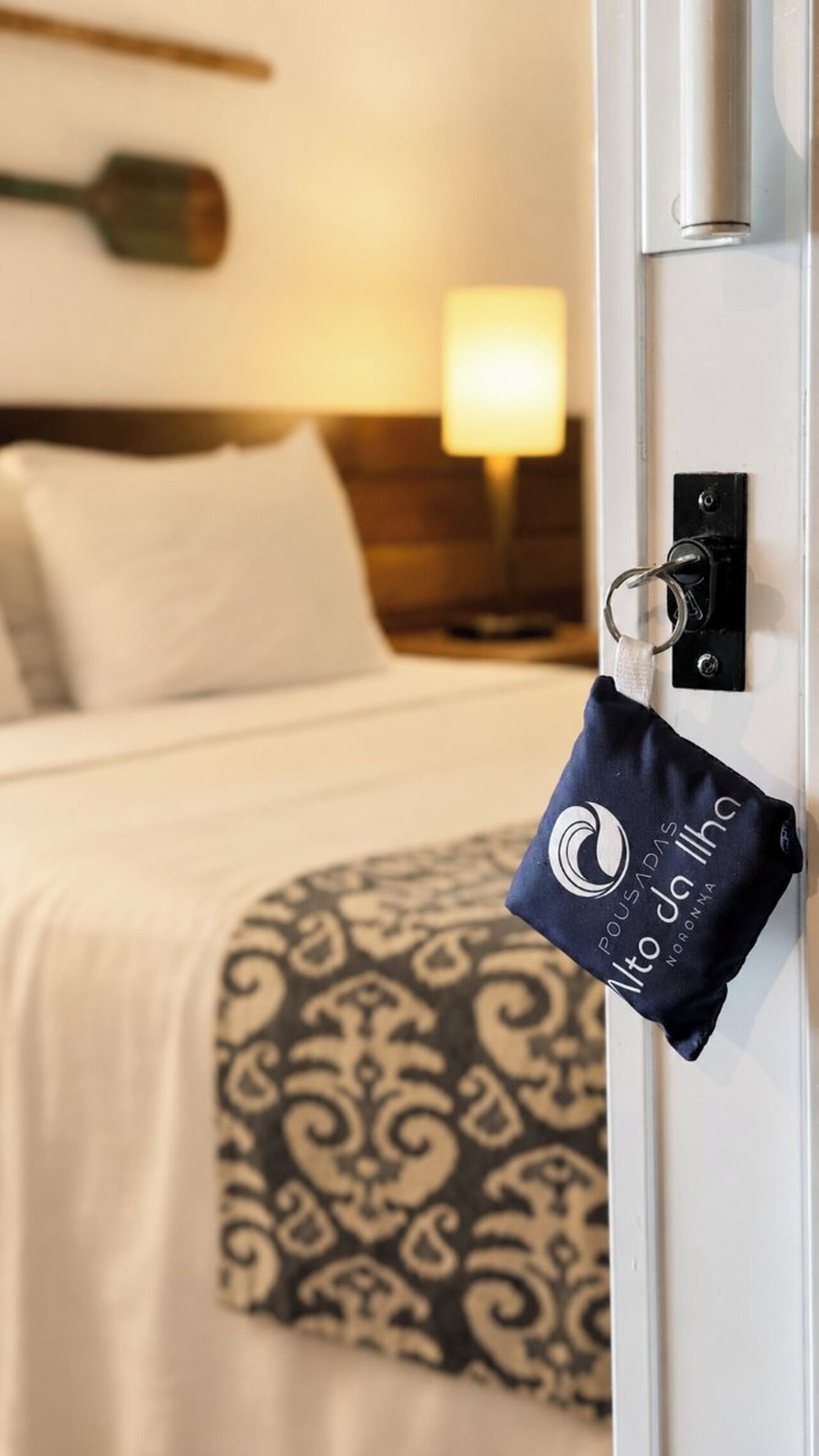 Quarto de hotel com cama, mesa de cabeceira, abajur e chaveiro com logotipo.