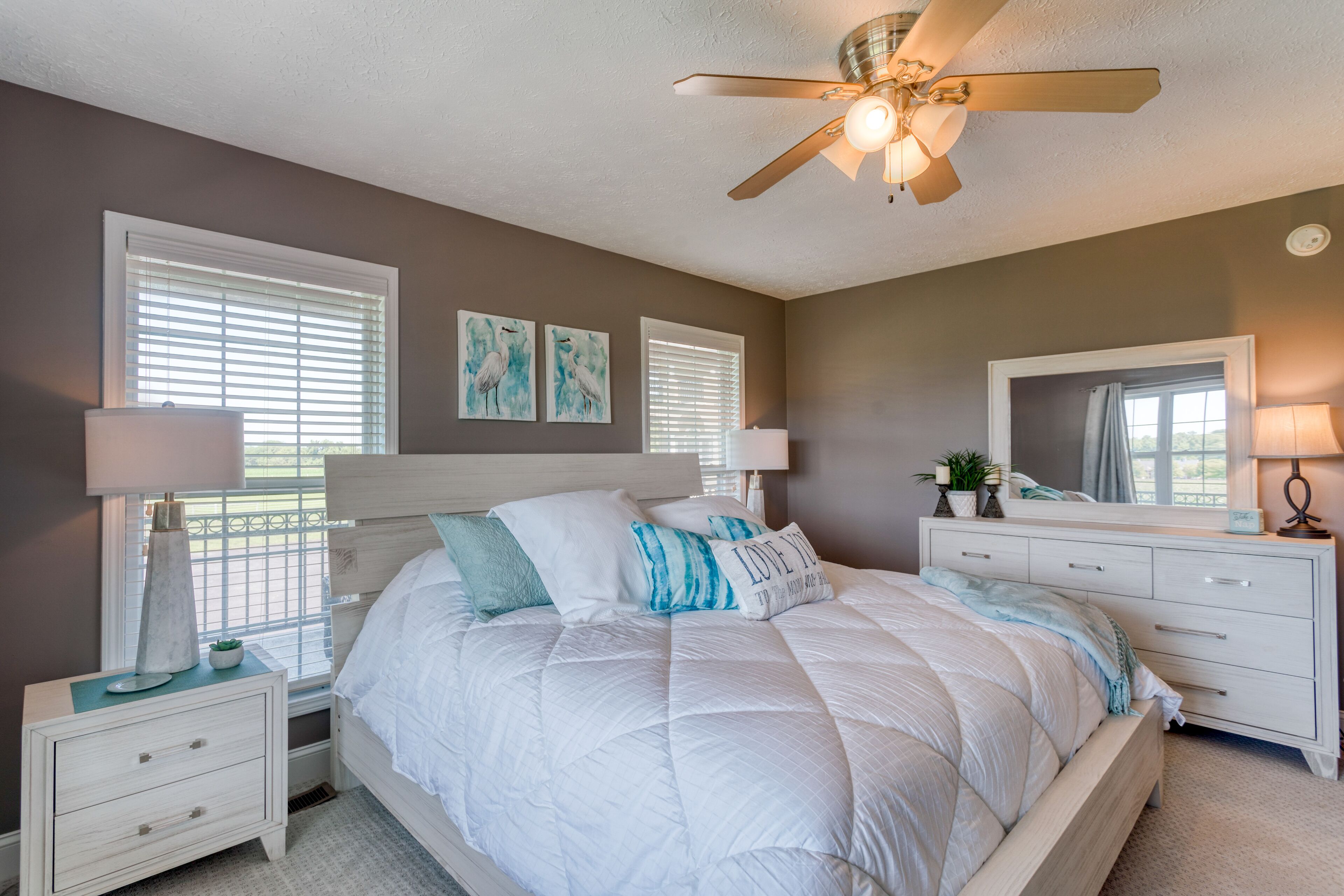 Une chambre à coucher comprenant un lit, des tables de chevet, une commode, un ventilateur de plafond et des œuvres d’art accrochées aux murs.
