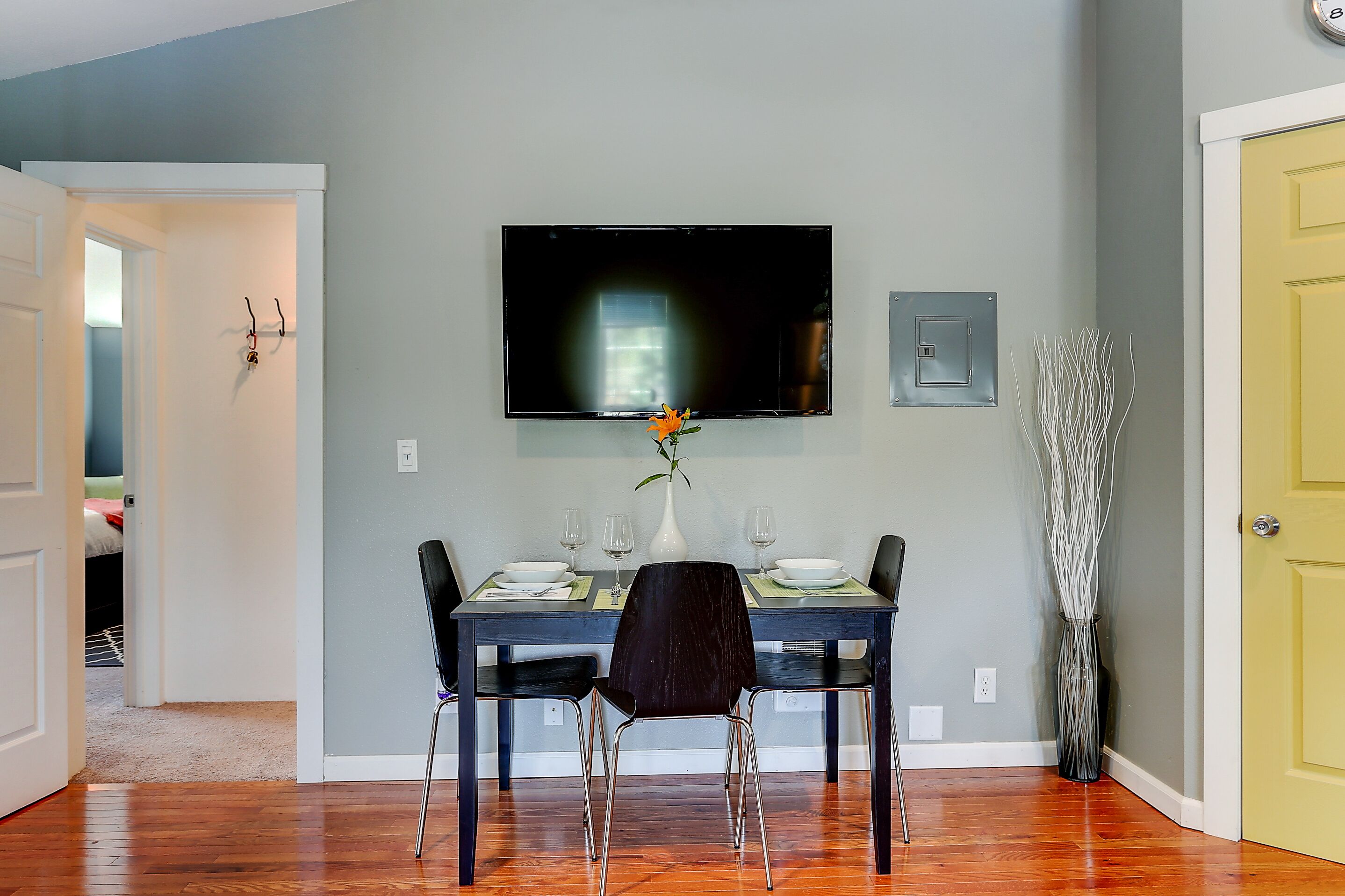 Un comedor con una mesa dispuesta para dos, un televisor de pantalla plana montado encima y un jarrón con una sola flor.