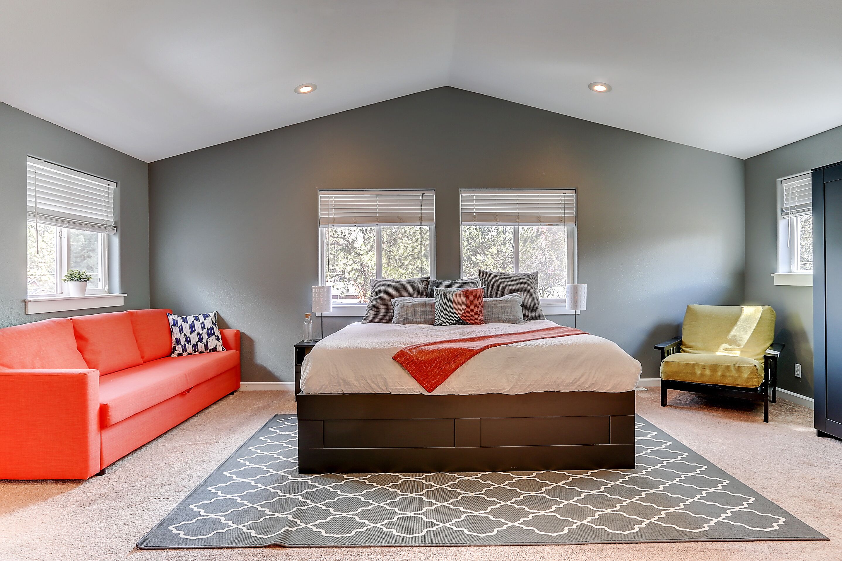 A bedroom with a large bed, a red sofa, a yellow armchair, and two windows with blinds.