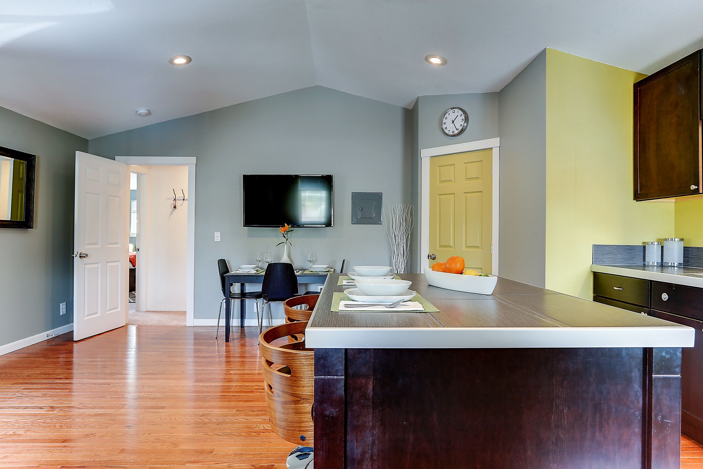 A modern kitchen with a large island, wooden chairs, a dining table, and a flat-screen TV mounted on the wall.