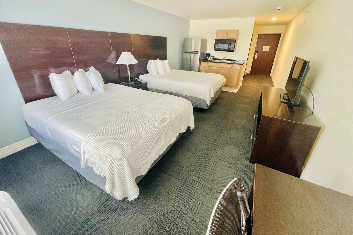 A hotel room with two beds, a desk, a TV, and a kitchenette.