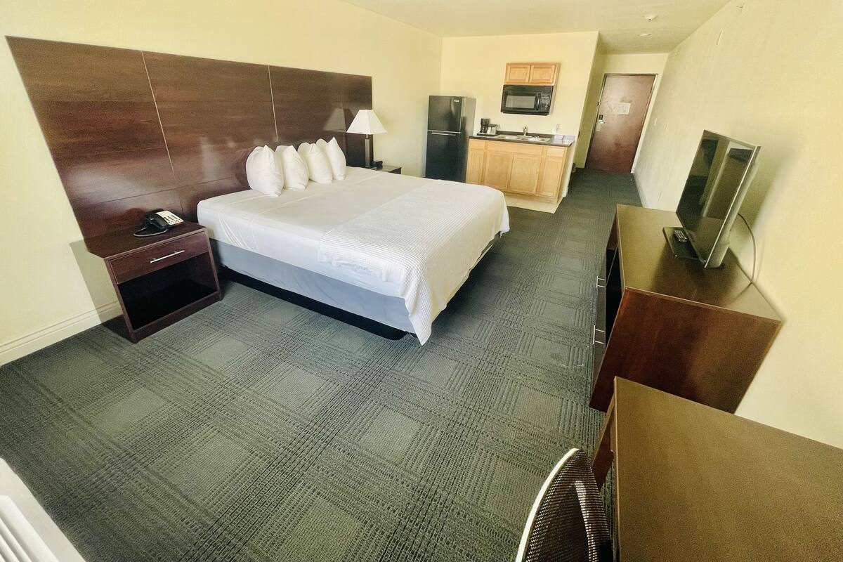 A hotel room with a bed, bedside table, lamp, TV, kitchenette, and a mirror.