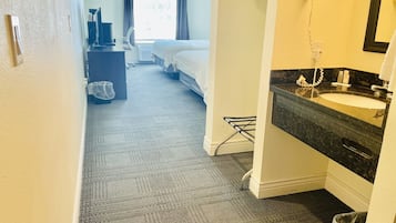 Una habitación de hotel con cama, escritorio con computadora, ventana con cortinas, baño con lavamanos y espejo, y piso alfombrado.