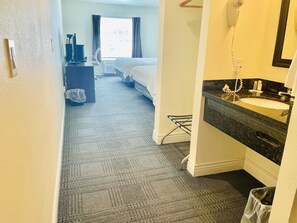 A hotel room with a bed, a desk with a computer, a window with curtains, a bathroom with a sink and mirror, and a carpeted floor.
