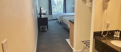 A hotel room with a carpeted floor, a desk with a lamp, a bed with a window, and a bathroom with a sink and a hairdryer.