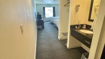 A hotel room with a bathroom featuring a sink, mirror, and hairdryer. A bed and a desk with a chair are visible in the background.