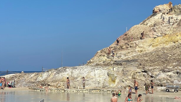 Una playa rocosa con una piscina, cerca hay una valla de madera y gente disfrutando en el agua.