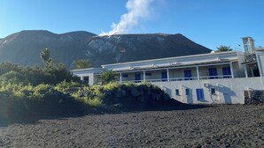Un edificio con fachada blanca y ventanas azules, ubicado en una playa con formaciones rocosas volcánicas al fondo.