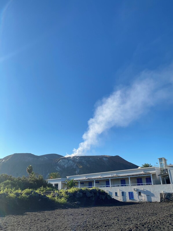 A building with a white facade and blue windows, situated on a beach with volcanic rock formations in the background.