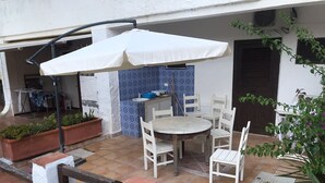 Un patio al aire libre con una mesa redonda y cuatro sillas, una sombrilla blanca y plantas en macetas.