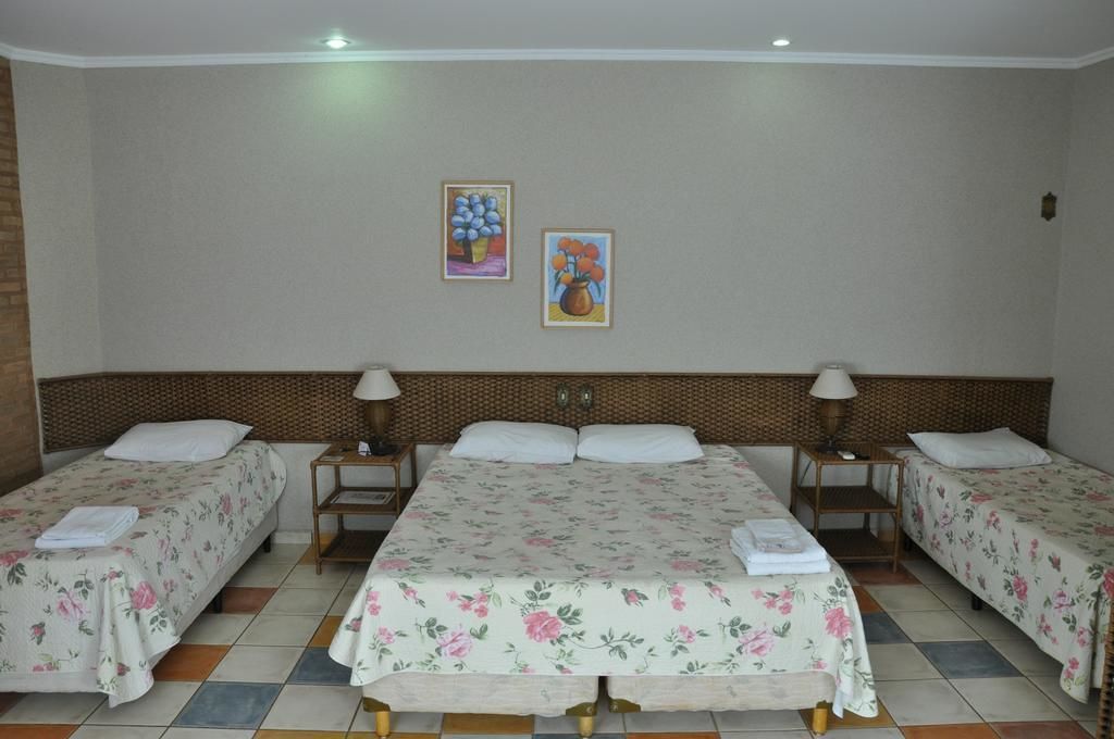 A hotel room with three single beds, each with a floral bedspread and white pillows. There are two bedside tables with lamps, and the room has a tiled floor.