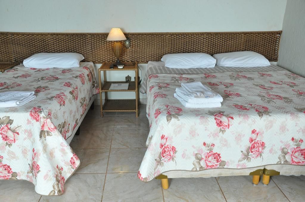 Une chambre d’hôtel avec deux lits simples, chacun agrémenté d’une couvre-lit à motifs floraux et de coussins blancs. Une petite table de chevet en bois avec une lampe se trouve entre les deux lits.