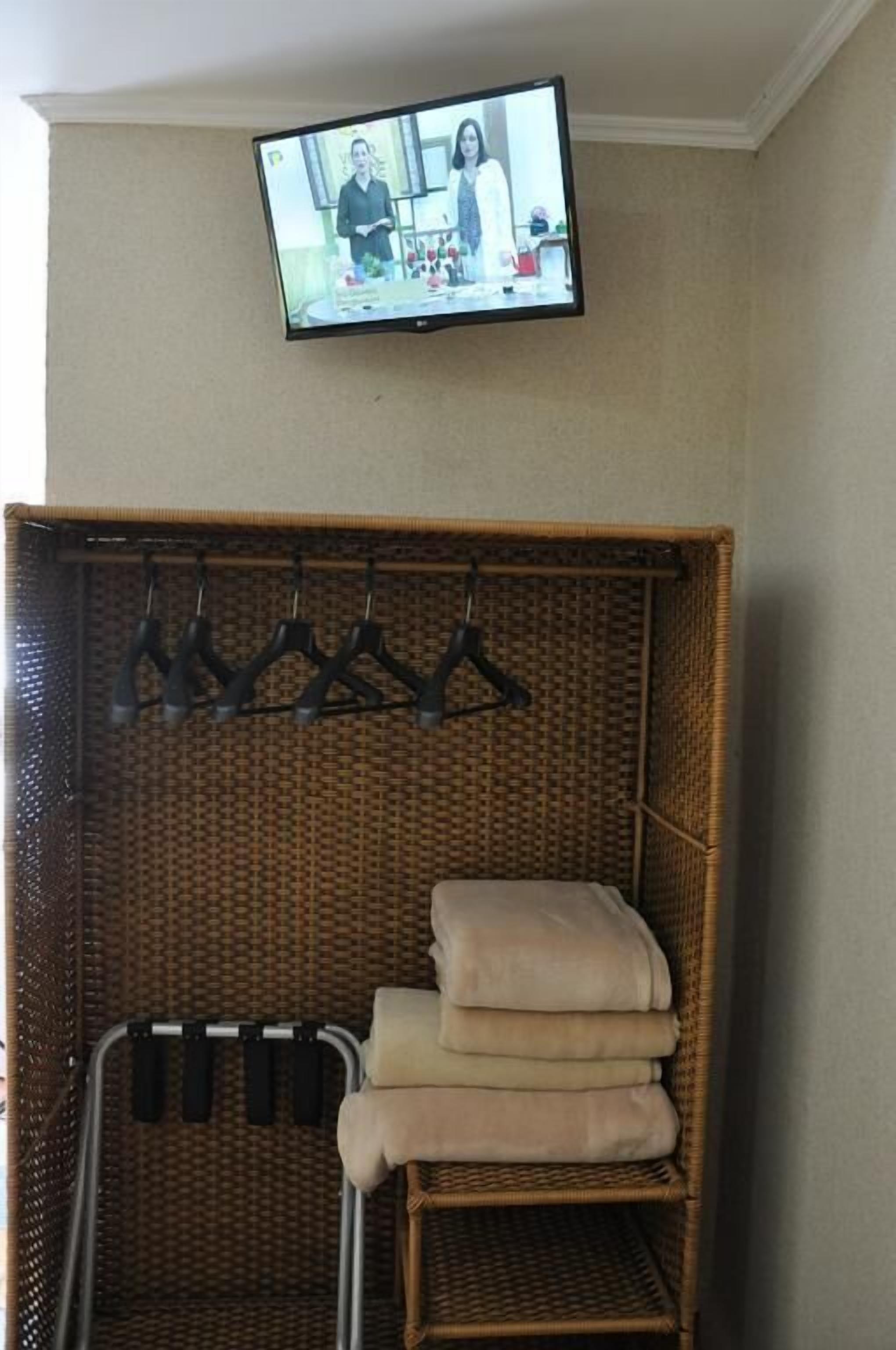 Ein Zimmer mit an der Wand montiertem Fernseher, der eine sprechende Person zeigt. Ein Korbstuhl mit gefalteter Decke und ein Kleiderständer mit Kleiderbügeln sind ebenfalls vorhanden.