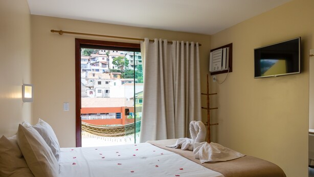 A hotel room with a bed, a window with curtains, a TV, and a view of buildings outside. - Costa Dourada Pousada (Arraial do Cabo)