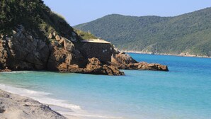A coastal scene with a rocky shoreline, clear turquoise water, and a building on a cliff. - Costa Dourada Pousada (Arraial do Cabo)