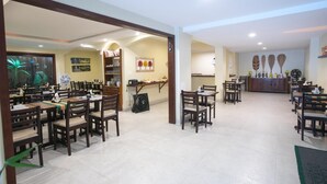 A spacious dining area with multiple wooden tables and chairs, a buffet counter, and a large window with a view of a plant. - Costa Dourada Pousada (Arraial do Cabo)