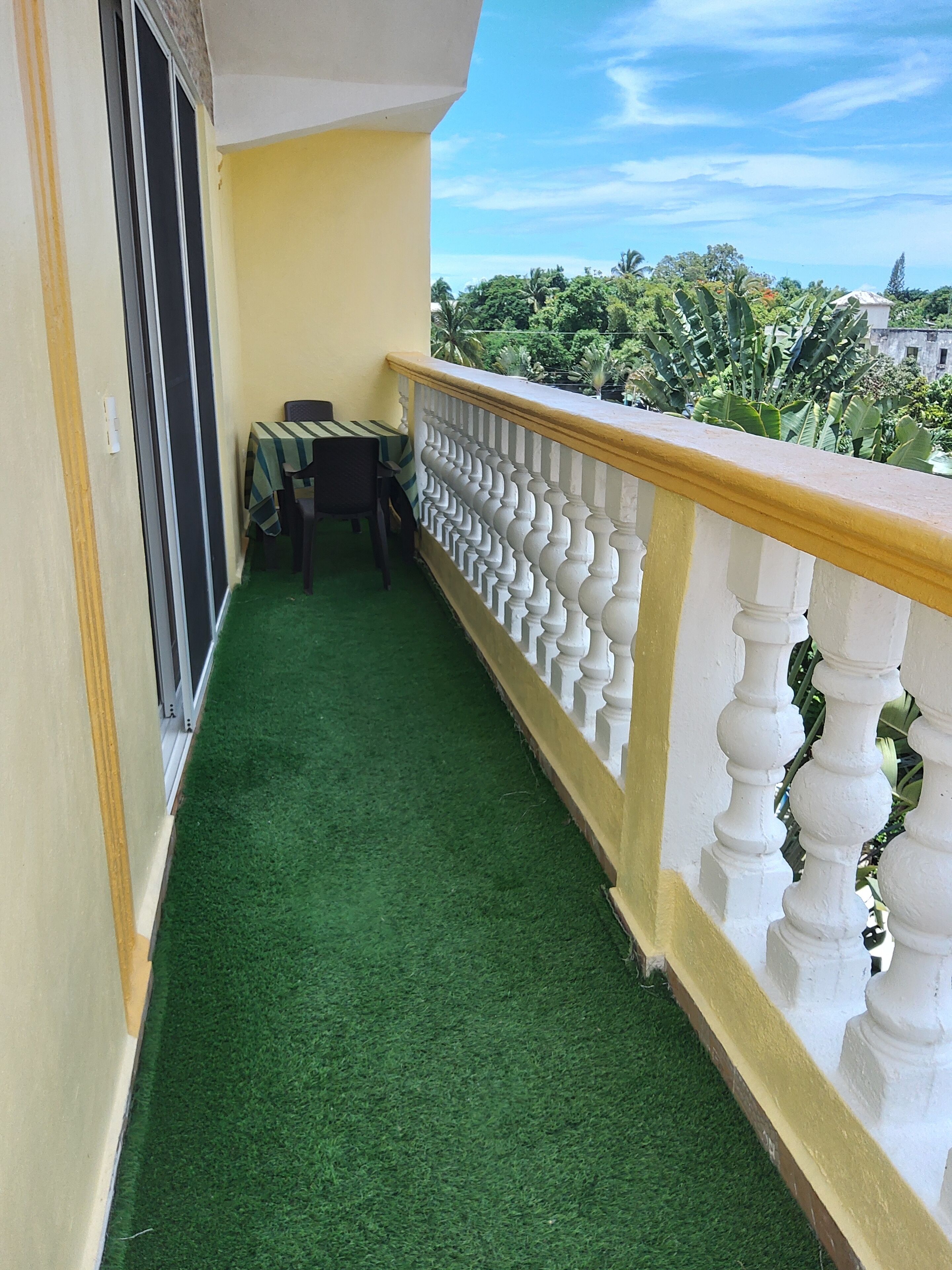 A balcony with artificial grass, a table with chairs, and a view of greenery and buildings.