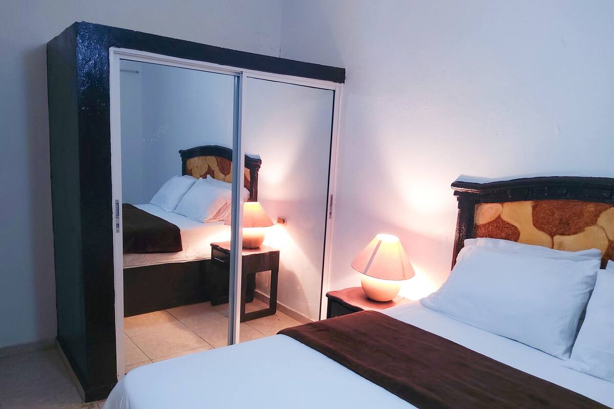 Hotellihuone, jossa on sänky, yöpöytä lampun kanssa, vaatekaappi ja peili.