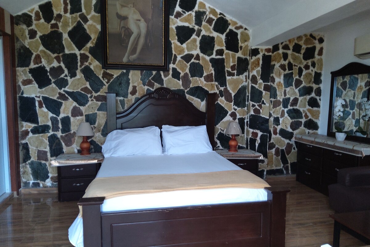 A bedroom with a stone wall, a wooden bed, two bedside tables with lamps, a dresser, and a mirror.
