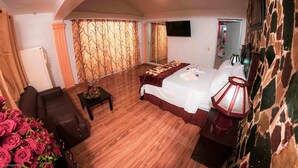 Ein Schlafzimmer mit Bett, Nachttisch, Lampe, Sofa, Tisch mit Blumen und Fernseher an der Wand.