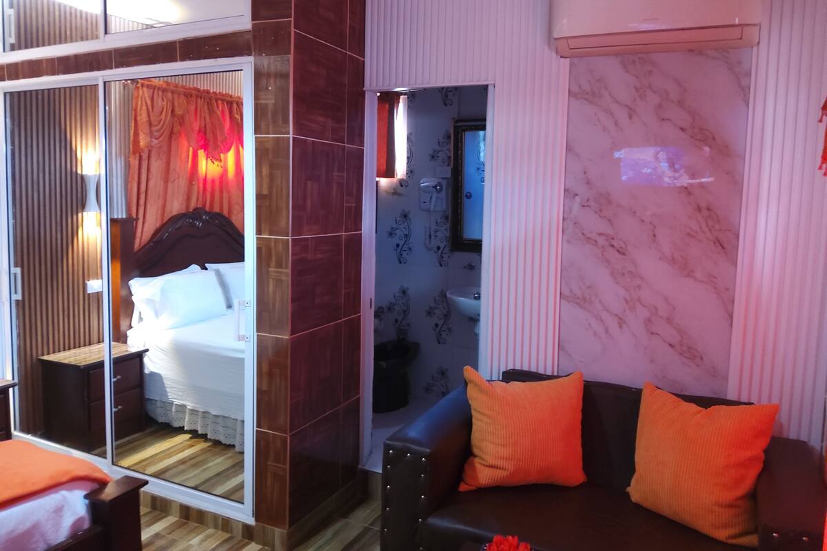 Hotellihuone, jossa on sänky, sohva, peili ja kylpyhuone, joka näkyy avoimesta ovesta.