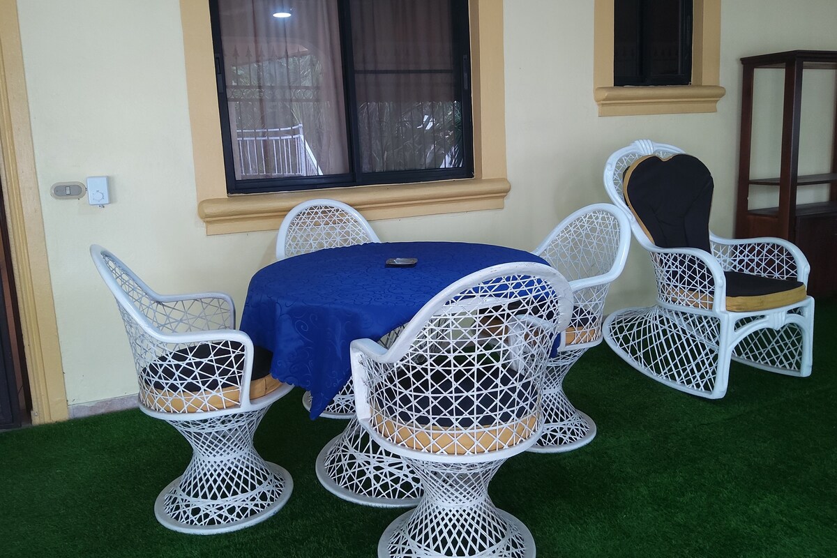 A dining area with a round table covered by a blue tablecloth, surrounded by white wicker chairs with cushioned seats.