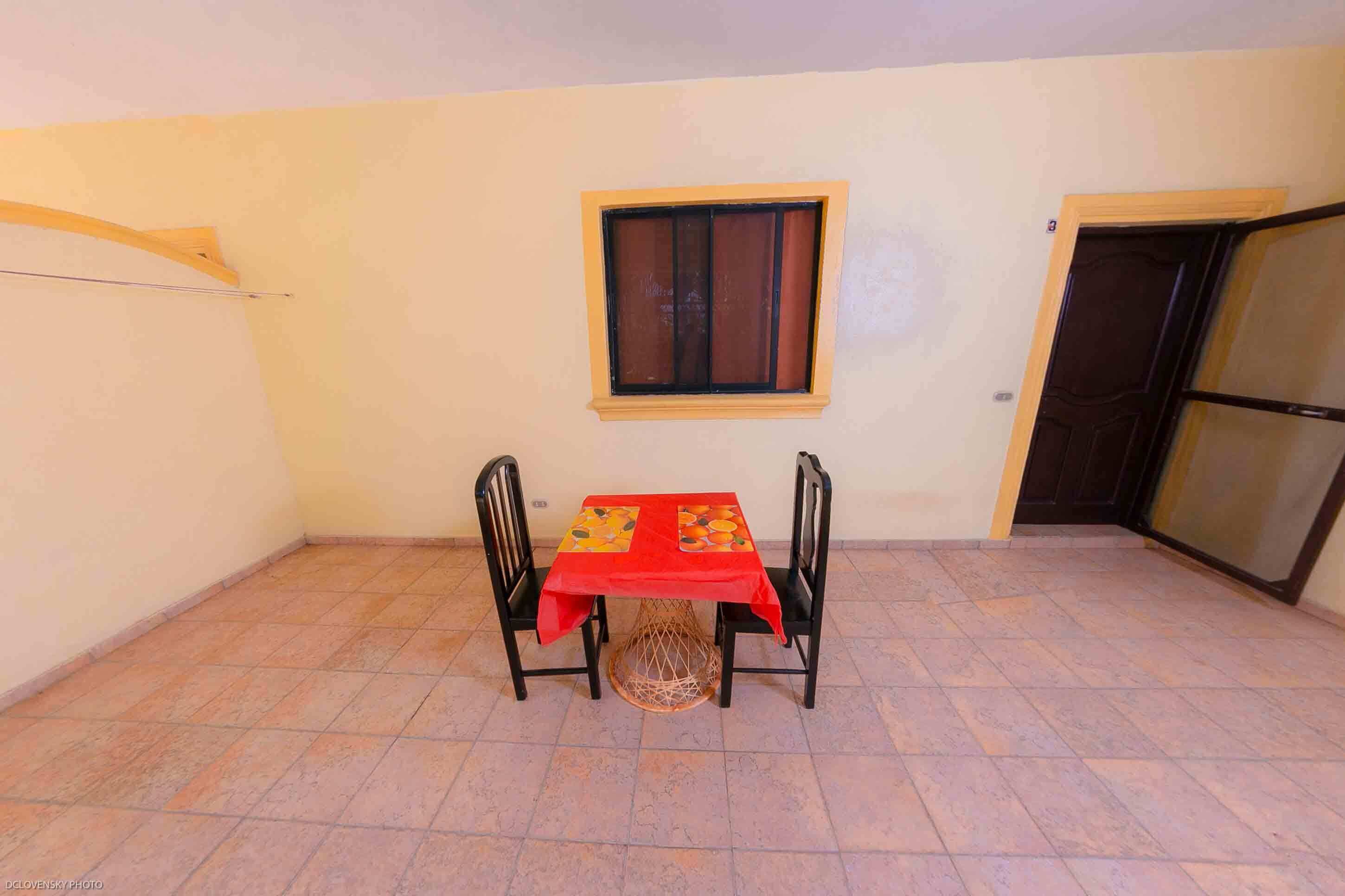 A small room with a tiled floor, a table with a red tablecloth, two chairs, a window with a brown frame, and a door with a glass panel.