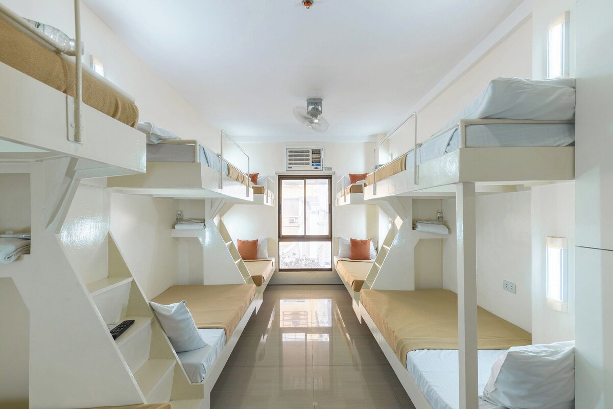 A dormitory room with bunk beds, a central window, and an air conditioning unit.