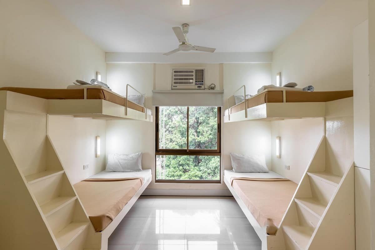 A dormitory room with bunk beds, a ceiling fan, an air conditioning unit, and a window with a view of trees.