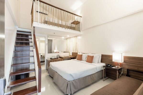 Quarto de hotel com uma cama, mesa de cabeceira, abajur e escadas que levam a um andar superior.