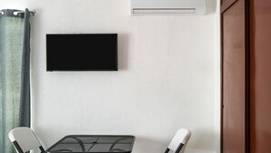 A room with a flat-screen TV mounted on the wall, a window with a curtain, and a table with two chairs.