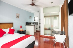 A hotel room with a bed, a desk, a chair, a kitchenette, and a sliding glass door to the balcony.