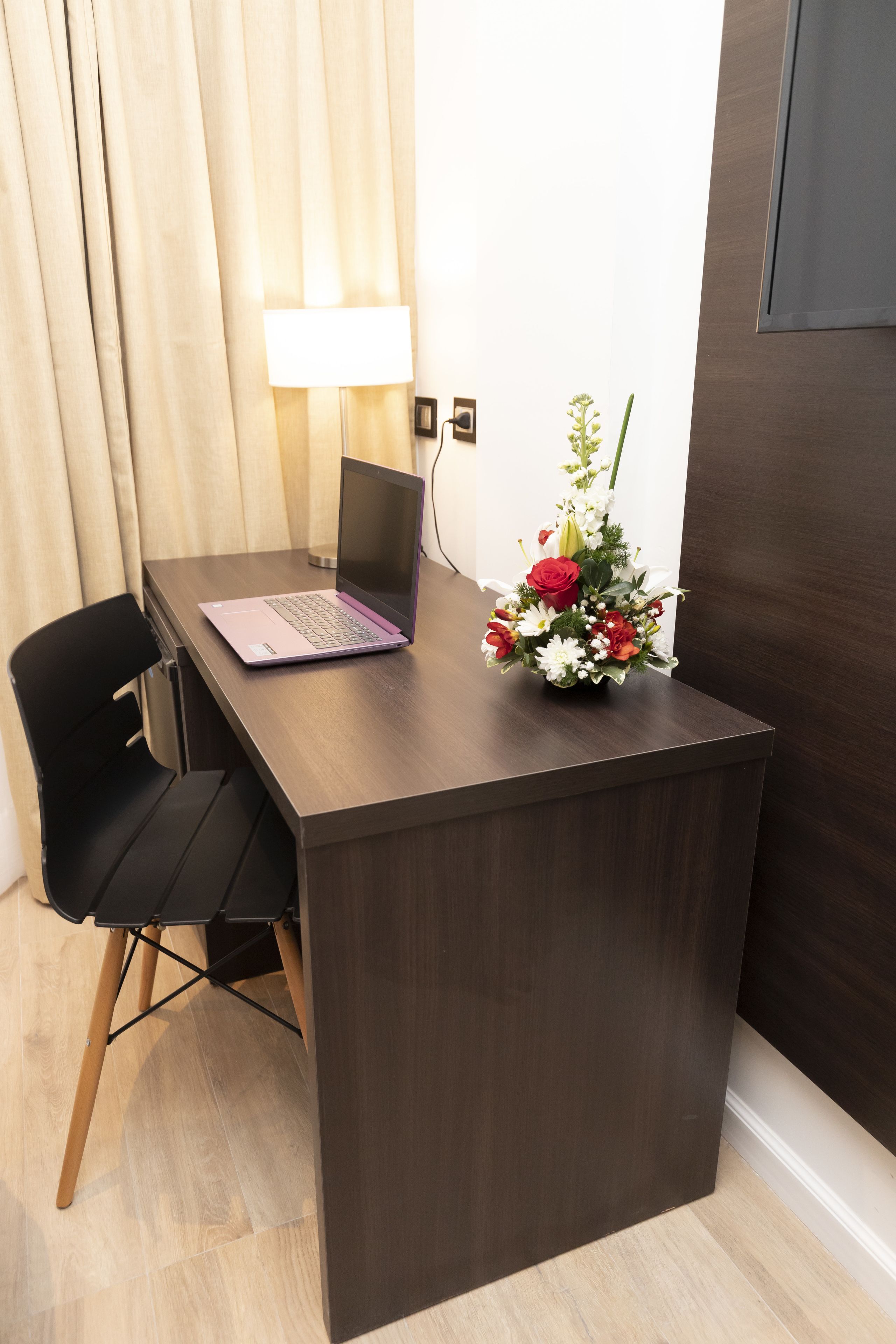 A wooden desk with a laptop, a black chair, a lamp, and a floral arrangement.