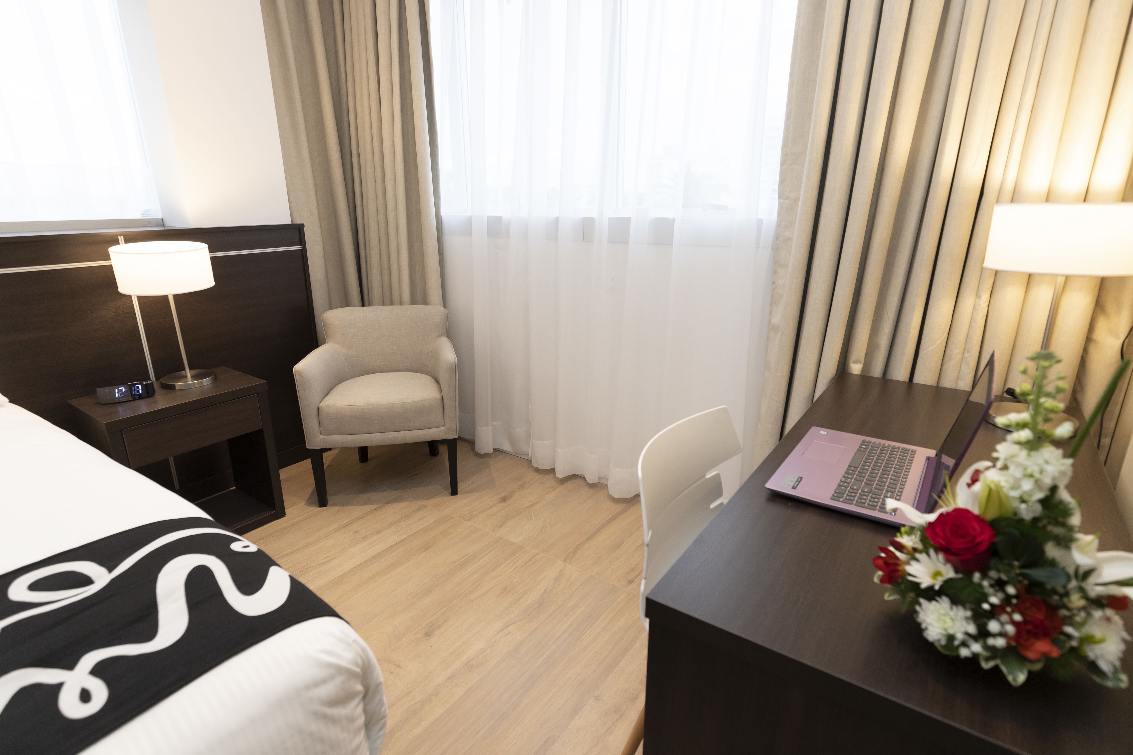 Ein Hotelzimmer mit einem Bett, einem Schreibtisch mit Laptop, einem Stuhl, einem Nachttisch mit Lampe und einem Fenster mit Vorhängen.