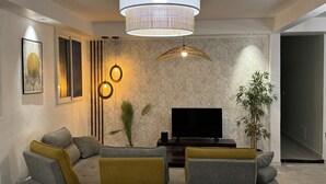 Un salon moderne avec un canapé d’angle, un téléviseur à écran plat, un éclairage décoratif et une grande fenêtre.