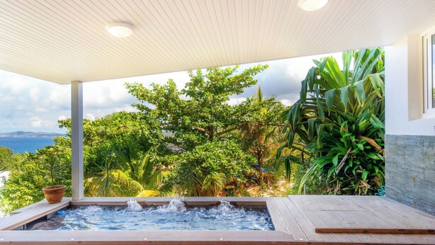 Un bain à remous avec une terrasse en bois, entouré d’une végétation luxuriante et offrant une vue sur la mer.