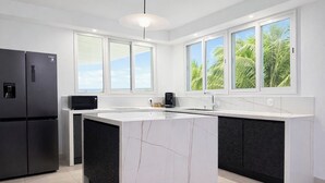 Une cuisine moderne dotée d’un îlot en marbre, de meubles de rangement noirs, d’un réfrigérateur noir, d’un four à micro-ondes et offrant une vue sur les palmiers à travers de grandes fenêtres.