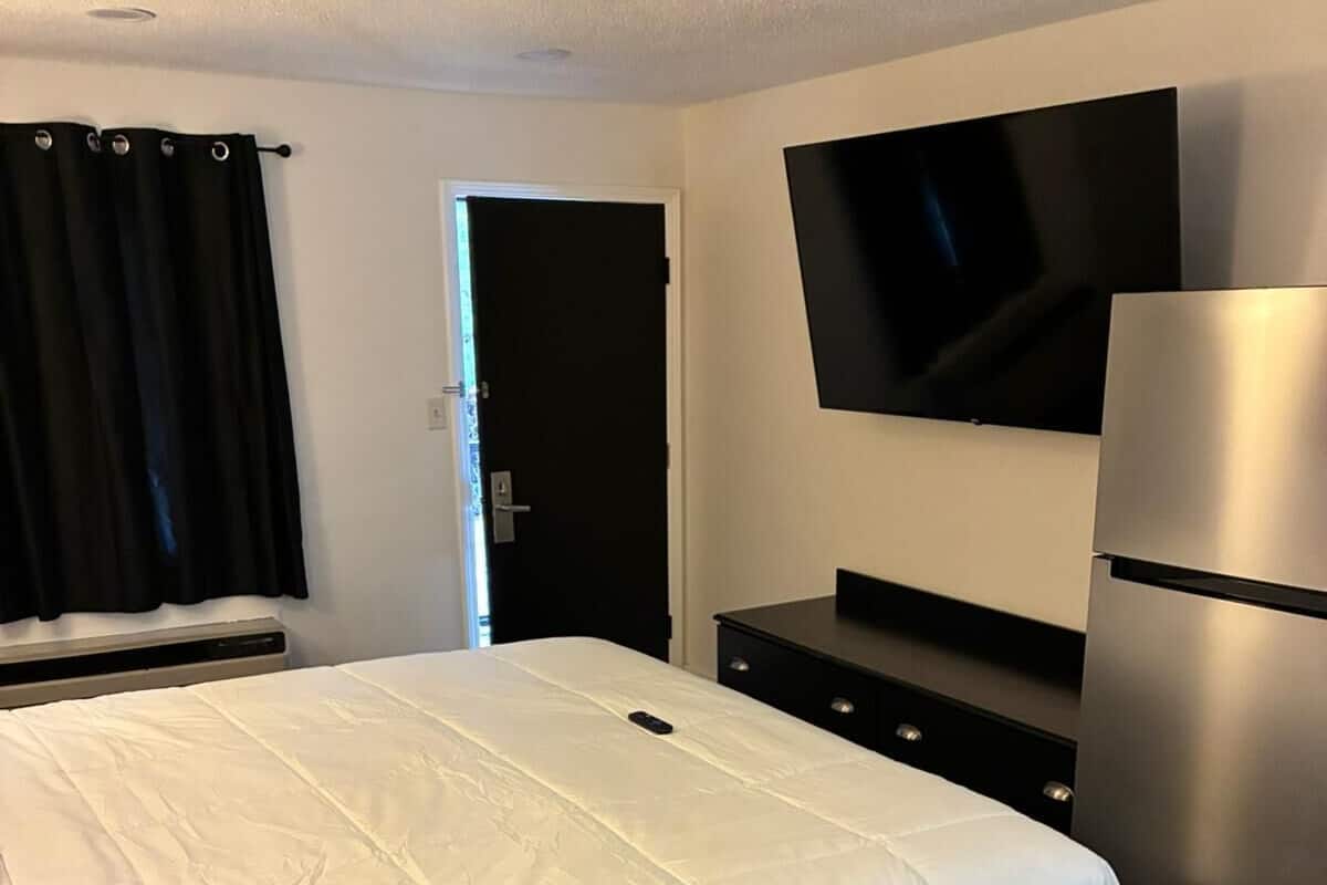 A hotel room with a bed, a flat-screen TV mounted on the wall, a refrigerator, and a dark-colored dresser.