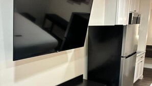 Moderni keittiö, jossa on seinälle asennettu taulutelevisio, musta työtaso vetolaatikoilla, ruostumattomasta teräksestä valmistettu jääkaappi ja valkoiset kaapit.